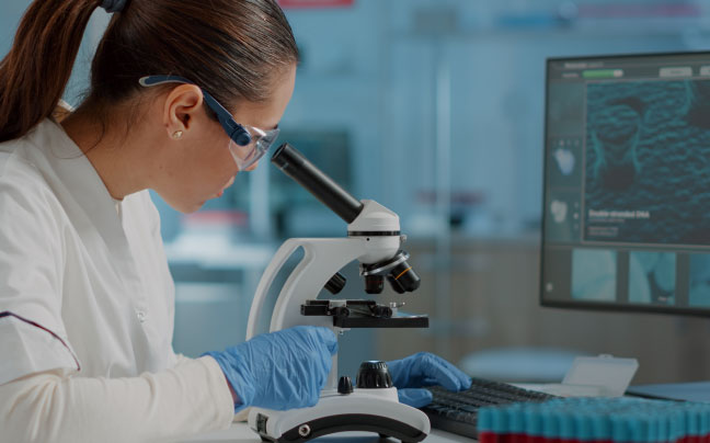Female scientist looking into a microscope with computer in the background.