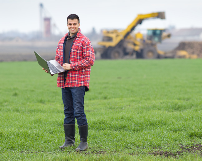 man standing in a field in front of heavy machinery holding laptop