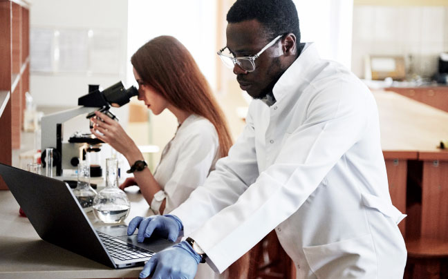 Female scientist looking into a microscope with computer in the background.