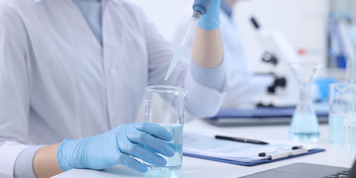 Scientist wearing blue gloves uses a pipette to transfer liquid into a beaker on a laboratory bench