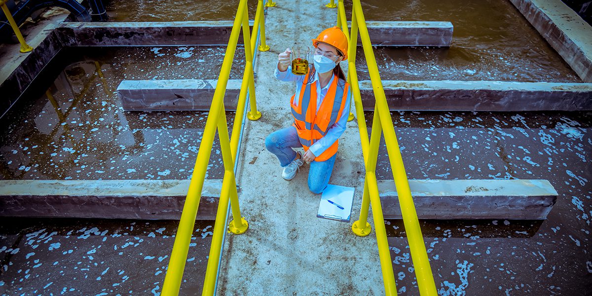 Construction worker in orange safety vest and hard hat kneels on a walkway beside a murky water tank, holding a sample container.