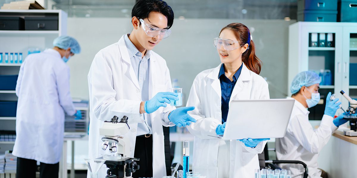 Two scientists in safety goggles and white coats discuss a sample in a modern lab with a microscope and laptop nearby.